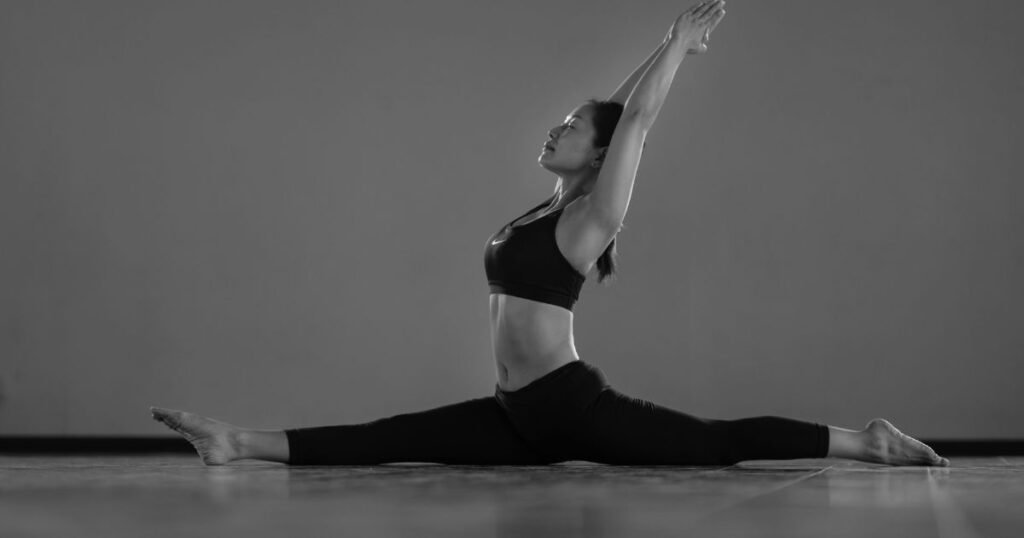 Woman performing a seated split stretch with both arms raised overhead on a studio floor.